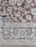 An intricately woven carpet with floral and geometric patterns. The design includes flowers, leaves, and symmetric motifs in blue and beige colors set against a brown background.