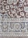 An intricately woven carpet with floral and geometric patterns. The design includes flowers, leaves, and symmetric motifs in blue and beige colors set against a brown background.