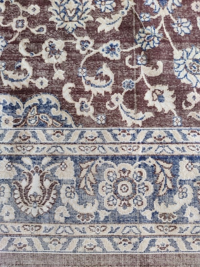 An intricately woven carpet with floral and geometric patterns. The design includes flowers, leaves, and symmetric motifs in blue and beige colors set against a brown background.