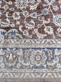 An intricately woven carpet with floral and geometric patterns. The design includes flowers, leaves, and symmetric motifs in blue and beige colors set against a brown background.