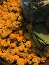 A pile of vibrant orange marigold flowers with some green leaves placed on top. The flowers are fresh and densely packed together, giving a lush and abundant appearance. On the top right, a transparent plastic bag containing green stems or foliage is visible.