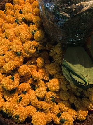 A pile of vibrant orange marigold flowers with some green leaves placed on top. The flowers are fresh and densely packed together, giving a lush and abundant appearance. On the top right, a transparent plastic bag containing green stems or foliage is visible.