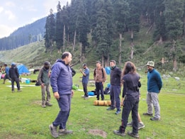 A group of people is gathered on a lush green lawn surrounded by tall pine trees. Some appear to be in conversation, while others are engaged in various activities. Camping gear and tents are visible, indicating a camp setup. The backdrop features a forested hillside, creating a serene outdoor setting.