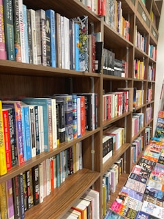 A bookstore shelf filled with a variety of books arranged in an organized manner. The books are of different genres, visible on multiple wooden shelves. There is a mix of hardcovers and paperbacks with colorful spines. Some books are placed upright, and others are stacked horizontally. Additionally, adjacent tables display more books laid out flat, offering a wide selection for browsing.