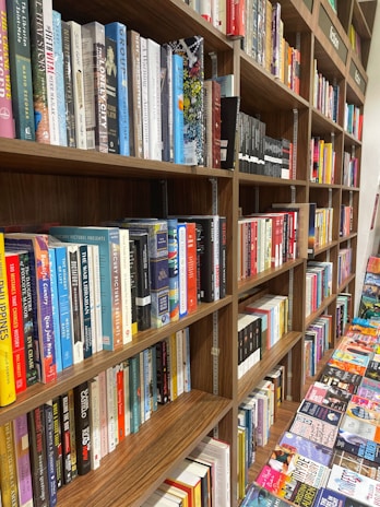 A bookstore shelf filled with a variety of books arranged in an organized manner. The books are of different genres, visible on multiple wooden shelves. There is a mix of hardcovers and paperbacks with colorful spines. Some books are placed upright, and others are stacked horizontally. Additionally, adjacent tables display more books laid out flat, offering a wide selection for browsing.