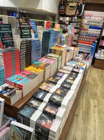 A well-organized bookstore display with stacks of colorful books placed on a wooden table. The books include popular titles with vibrant covers. Surrounding shelves hold an assortment of stationery items and decorative products. The setting appears bright and inviting, with neatly arranged items enhancing the visual appeal.