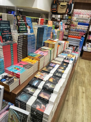 A well-organized bookstore display with stacks of colorful books placed on a wooden table. The books include popular titles with vibrant covers. Surrounding shelves hold an assortment of stationery items and decorative products. The setting appears bright and inviting, with neatly arranged items enhancing the visual appeal.