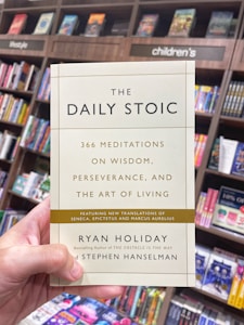 A hand is holding a book titled 'The Daily Stoic' in front of a bookshelf filled with various books. The book cover mentions 366 meditations on wisdom, perseverance, and the art of living, featuring new translations of Seneca, Epictetus, and Marcus Aurelius. The authors are Ryan Holiday and Stephen Hanselman. The surrounding bookshelf sections are labeled with categories such as lifestyle and children's.