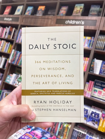 A hand is holding a book titled 'The Daily Stoic' in front of a bookshelf filled with various books. The book cover mentions 366 meditations on wisdom, perseverance, and the art of living, featuring new translations of Seneca, Epictetus, and Marcus Aurelius. The authors are Ryan Holiday and Stephen Hanselman. The surrounding bookshelf sections are labeled with categories such as lifestyle and children's.