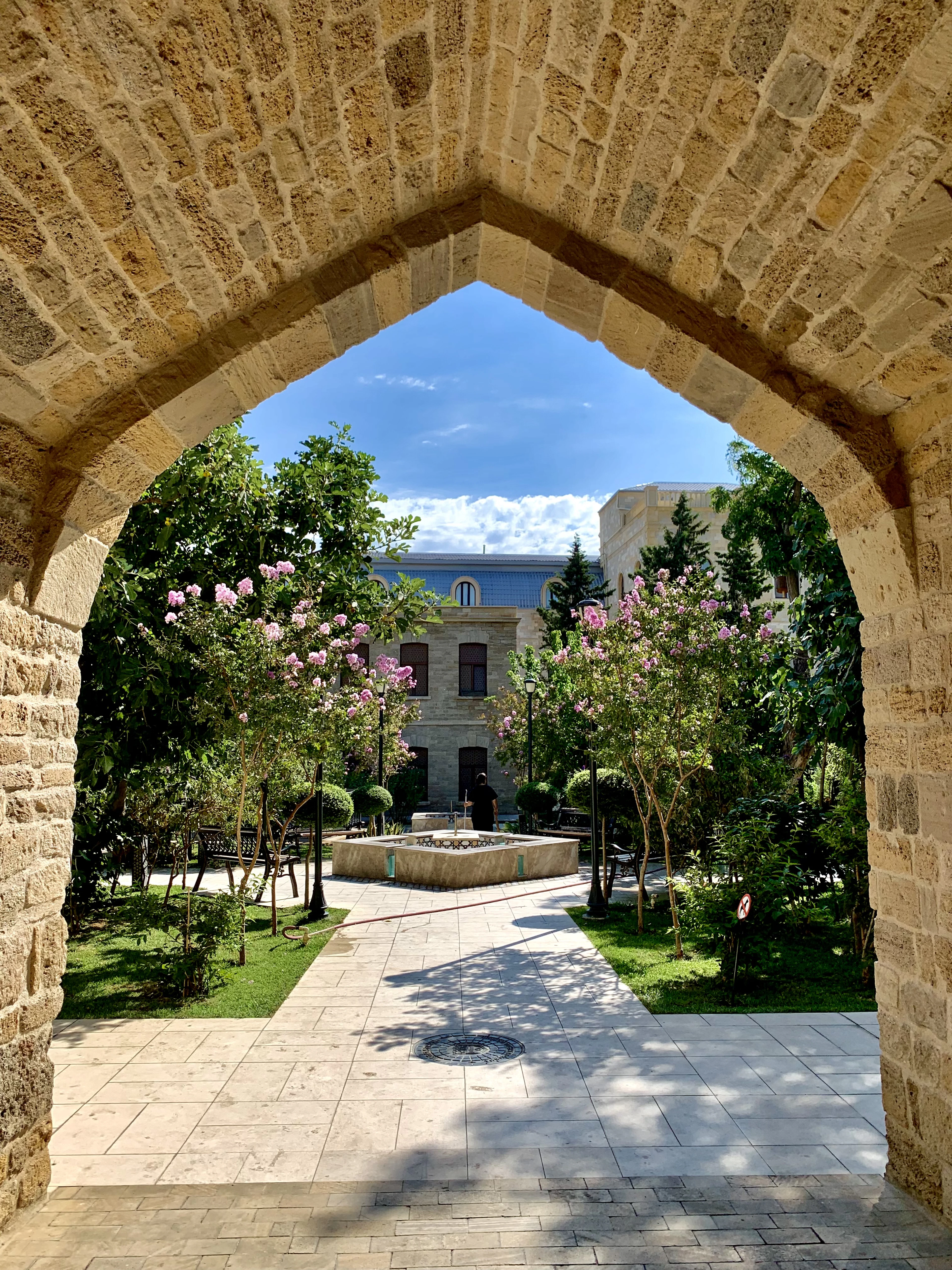 A stone archway frames a quiet courtyard with blossoming trees and a small fountain in Baku's Old City.