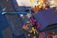 A rooftop scattered with orange and yellow autumn leaves. A red-handled hammer, a blue crowbar, and a pair of yellow-handled pliers rest on the shingles. A rectangular vent cover is visible among the leaves.