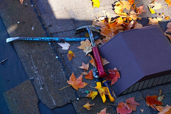 A rooftop scattered with orange and yellow autumn leaves. A red-handled hammer, a blue crowbar, and a pair of yellow-handled pliers rest on the shingles. A rectangular vent cover is visible among the leaves.