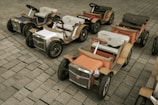 Several vintage-style toy cars are neatly arranged on a paved surface. The cars are colorful with chrome accents, wooden frames, and intricate detailing, each featuring a steering wheel and cushioned seat.