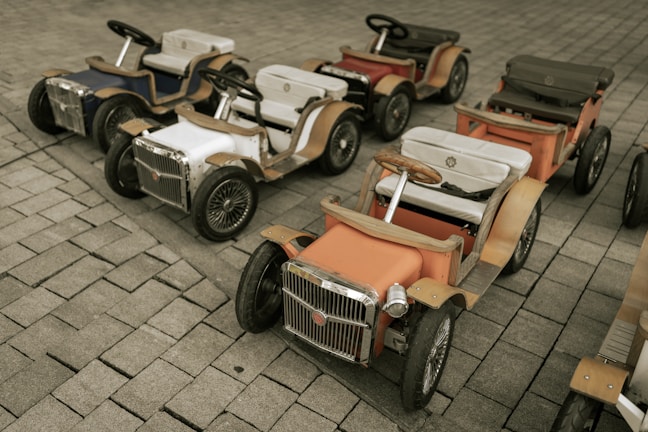 Several vintage-style toy cars are neatly arranged on a paved surface. The cars are colorful with chrome accents, wooden frames, and intricate detailing, each featuring a steering wheel and cushioned seat.