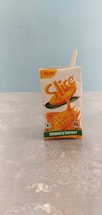A small tetra pack of mango juice labeled 'Slice' is placed upright on a floral patterned surface. The pack is primarily white with orange and green accents, featuring an image of a mango. A drinking straw is attached to the side, and the price of ₹10.00 is marked at the top.