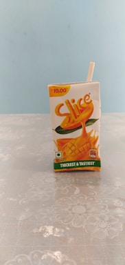 A small tetra pack of mango juice labeled 'Slice' is placed upright on a floral patterned surface. The pack is primarily white with orange and green accents, featuring an image of a mango. A drinking straw is attached to the side, and the price of ₹10.00 is marked at the top.