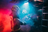 A firefighter wearing protective gear is surrounded by dramatic red and blue lighting, creating a moody and intense atmosphere. The figure is holding a tool, possibly engaging with equipment amidst swirling smoke.