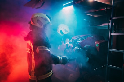 A firefighter wearing protective gear is surrounded by dramatic red and blue lighting, creating a moody and intense atmosphere. The figure is holding a tool, possibly engaging with equipment amidst swirling smoke.