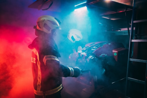 A firefighter wearing protective gear is surrounded by dramatic red and blue lighting, creating a moody and intense atmosphere. The figure is holding a tool, possibly engaging with equipment amidst swirling smoke.