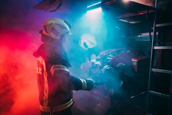 A firefighter wearing protective gear is surrounded by dramatic red and blue lighting, creating a moody and intense atmosphere. The figure is holding a tool, possibly engaging with equipment amidst swirling smoke.