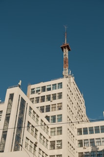 A multi-story white building with a grid of windows rises into a clear blue sky, topped with a tall communication tower featuring multiple antennas.