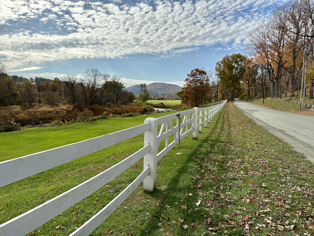 A white rail fence along a road near Woodstock, Vermont, with autumn trees and a hill behind