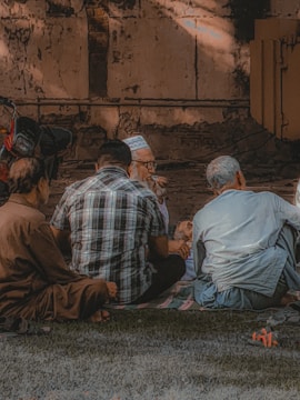 Several individuals are sitting together on a grassy area, engaging in conversation. They are positioned in a semicircle facing each other. The background features an old, textured wall that appears weathered. Light and shadow interplay on the walls and the ground, adding depth to the scene.