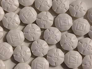 A close-up view of numerous white, round, tablet-like pills with markings '30' and 'M' embossed on them. The pills are arranged in a scattered pattern filling the entire frame.