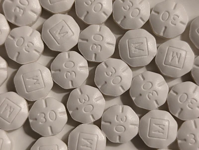 A close-up view of numerous white, round, tablet-like pills with markings '30' and 'M' embossed on them. The pills are arranged in a scattered pattern filling the entire frame.