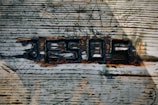 A textured wooden surface with the word 'JESUS' carved into it. The letters are slightly darkened, with burnt or charred edges, making them stand out against the lighter wood grain. Various geometric shapes are etched above the word, adding an abstract touch. The play of light and shadow across the wood enhances the depth and roughness of the surface.