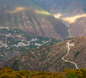 A winding mountain path through the Atlas Mountains with lush greenery and a small Berber village