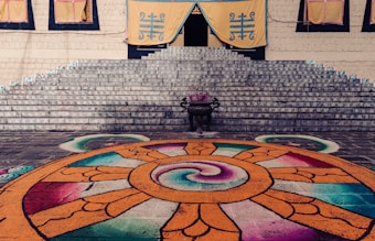 A set of stone steps leads up to a building adorned with yellow banners featuring blue symbols. In front of the steps is a vibrant, colorful mandala design on the ground with intricate patterns in orange, red, purple, and teal. A black incense burner is placed at the base of the steps, adding a traditional and ceremonial touch to the scene.
