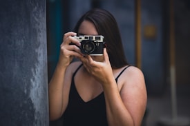A person holds a vintage camera up to their face, focusing on taking a photograph. The background is softly blurred, with muted tones adding depth. The person's long hair and sleeveless attire suggest a casual setting.