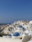 A picturesque coastal town features whitewashed buildings with iconic blue domes scattered across a hillside. These structures overlook the deep blue sea under a clear sky. The architecture reflects traditional Cycladic design, and there are hints of vegetation, including a few palm trees.