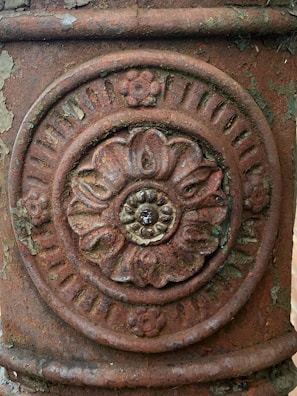 An intricately carved floral pattern is embossed on a weathered, rust-colored metallic surface. The design features a central flower with petals radiating outward, surrounded by a circular border with smaller decorative elements. The surface shows signs of aging and peeling paint, adding texture and character.