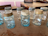 A collection of vintage mason jars with varying shades of blue and clear glass arranged on a rustic wooden table. Some jars have metal lids in different states of rust and wear, while others are lidless. The background consists of additional wooden furniture and pale pink wall panels.