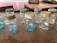 A collection of vintage mason jars with varying shades of blue and clear glass arranged on a rustic wooden table. Some jars have metal lids in different states of rust and wear, while others are lidless. The background consists of additional wooden furniture and pale pink wall panels.
