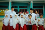 A group of children and two adults are gathered together, wearing uniforms with white tops and red bottoms. The children appear to be school students, some smiling, with a couple wearing face masks. The adults, wearing dark hats, stand behind the children with cheerful expressions, raising their thumbs up. They are in front of a blue building with large windows.
