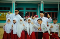 A group of children and two adults are gathered together, wearing uniforms with white tops and red bottoms. The children appear to be school students, some smiling, with a couple wearing face masks. The adults, wearing dark hats, stand behind the children with cheerful expressions, raising their thumbs up. They are in front of a blue building with large windows.
