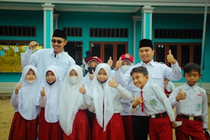 A group of children and two adults are gathered together, wearing uniforms with white tops and red bottoms. The children appear to be school students, some smiling, with a couple wearing face masks. The adults, wearing dark hats, stand behind the children with cheerful expressions, raising their thumbs up. They are in front of a blue building with large windows.