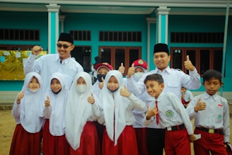 A group of children and two adults are gathered together, wearing uniforms with white tops and red bottoms. The children appear to be school students, some smiling, with a couple wearing face masks. The adults, wearing dark hats, stand behind the children with cheerful expressions, raising their thumbs up. They are in front of a blue building with large windows.