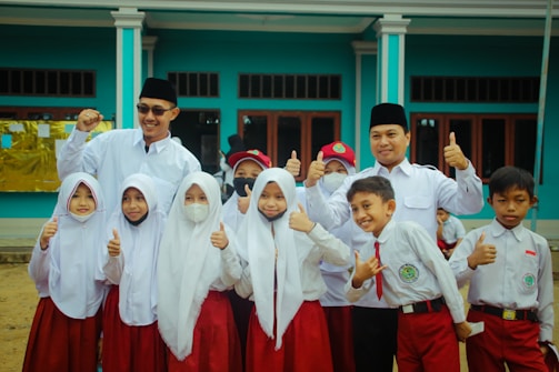 A group of children and two adults are gathered together, wearing uniforms with white tops and red bottoms. The children appear to be school students, some smiling, with a couple wearing face masks. The adults, wearing dark hats, stand behind the children with cheerful expressions, raising their thumbs up. They are in front of a blue building with large windows.
