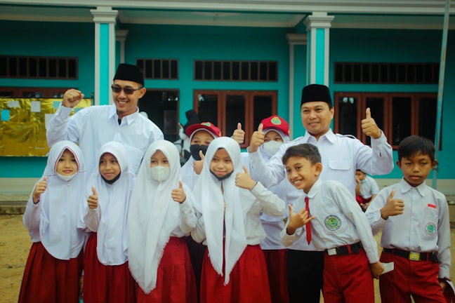 A group of children and two adults are gathered together, wearing uniforms with white tops and red bottoms. The children appear to be school students, some smiling, with a couple wearing face masks. The adults, wearing dark hats, stand behind the children with cheerful expressions, raising their thumbs up. They are in front of a blue building with large windows.