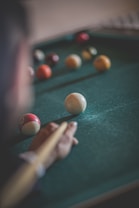 A close-up view of a billiard table with several balls scattered across its surface. A hand is holding a cue stick, aiming at a white ball in the foreground.
