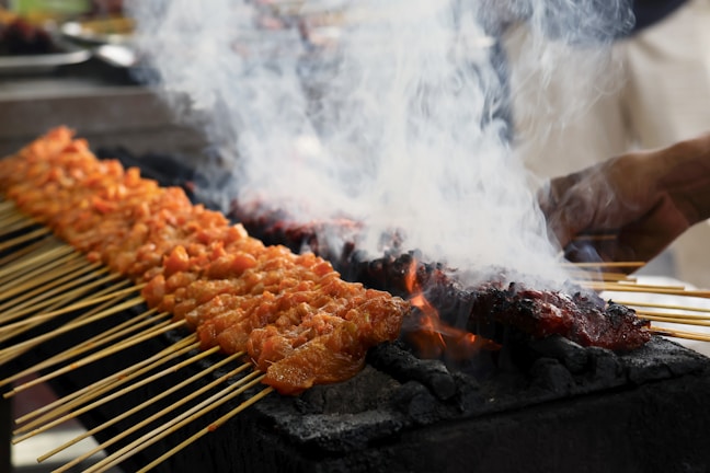 Skewers of marinated meat are being grilled over charcoal, with white smoke rising from the hot coals. The skewers are neatly arranged in rows, and a hand is visible tending to the grill.