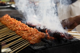 Skewers of marinated meat are being grilled over charcoal, with white smoke rising from the hot coals. The skewers are neatly arranged in rows, and a hand is visible tending to the grill.