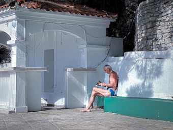 An elderly man wearing blue swim trunks is sitting alone on a painted concrete structure. He is outdoors, in front of a white building with a red-tiled roof and stone walls, reading a book or a magazine.