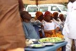 A group of people are seated under a canopy, with several women dressed in white tops and vibrant orange and yellow patterned headwraps and skirts. A man in a blue outfit is also seated among them. In the foreground, there are plates with food on a table, and a white vehicle is visible in the background.