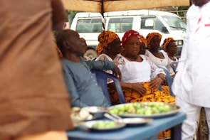 A group of people are seated under a canopy, with several women dressed in white tops and vibrant orange and yellow patterned headwraps and skirts. A man in a blue outfit is also seated among them. In the foreground, there are plates with food on a table, and a white vehicle is visible in the background.