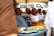 A group of people are seated under a canopy, with several women dressed in white tops and vibrant orange and yellow patterned headwraps and skirts. A man in a blue outfit is also seated among them. In the foreground, there are plates with food on a table, and a white vehicle is visible in the background.